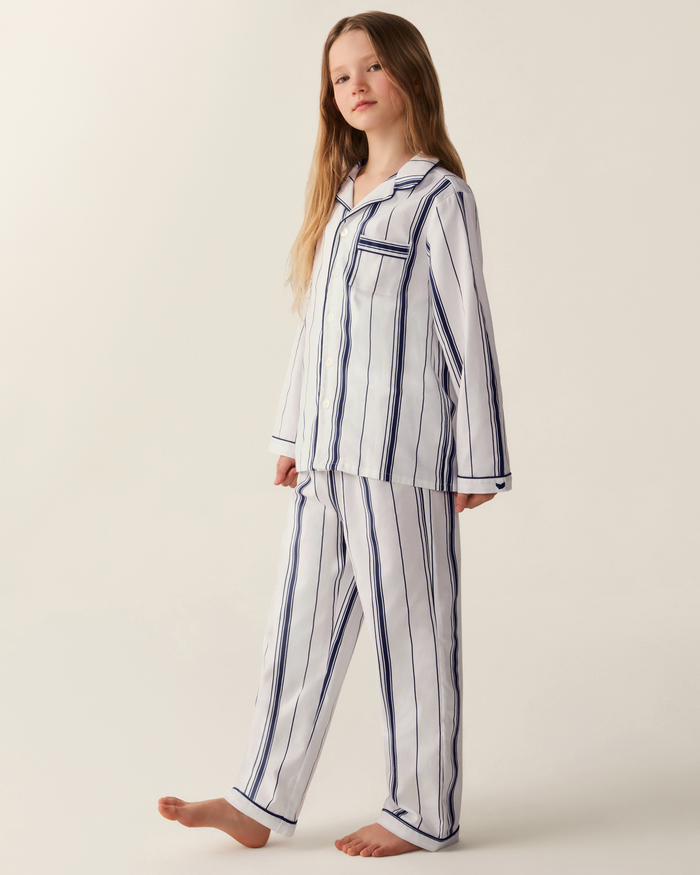 A young girl stands barefoot against a plain light background, dressed in the Kid’s Twill Pajama Set in Parlor Stripe, which features a collared long-sleeve top and matching white pajamas with navy blue vertical stripes.