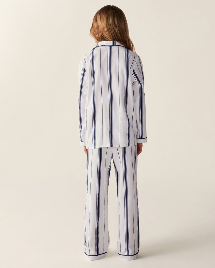 A child with long light brown hair is seen from behind, wearing the Kid’s Twill Pajama Set in Parlor Stripe—white pajamas featuring vertical navy blue stripes—against a plain light background.