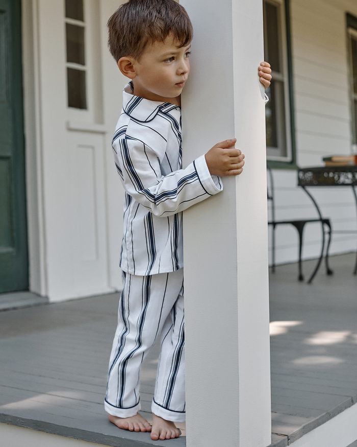 A young boy, barefoot and looking to the side, stands on a porch holding a white pillar while wearing the Kid’s Twill Pajama Set in Parlor Stripe. The background features a door, window, and outdoor furniture.