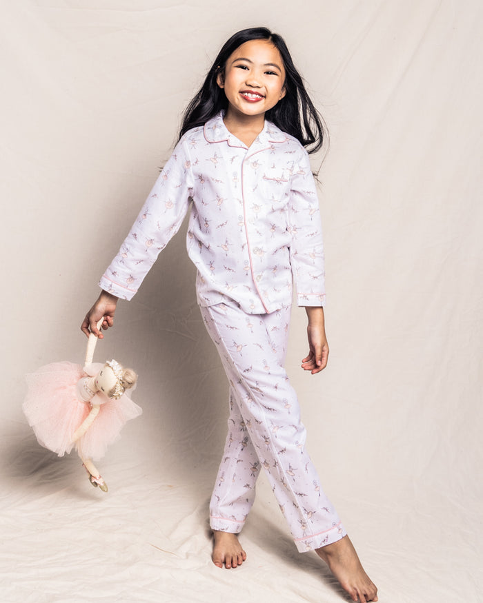 A young girl with long, dark hair smiles while holding a plush doll. Shes wearing Petite Plumes Kids Twill Pajama Set in Sugar Plum Fairy, made from the finest quality cotton, and stands barefoot against a light background.