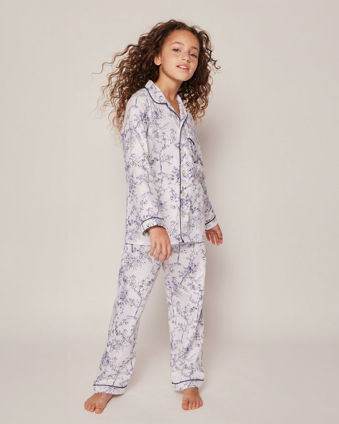 The Kids Twill Pajama Set in Timeless Toile by Petite Plume features a long-sleeve, luxury sleepwear style with a classic collar and matching pants. Its white background showcases an intricate blue floral and bird pattern for a vintage, sophisticated charm.