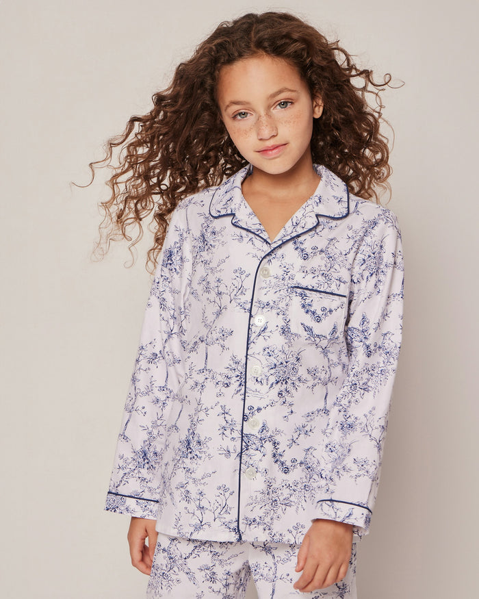 The Kids Twill Pajama Set in Timeless Toile by Petite Plume features a long-sleeve, luxury sleepwear style with a classic collar and matching pants. Its white background showcases an intricate blue floral and bird pattern for a vintage, sophisticated charm.