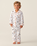 A young child with light brown hair stands barefoot, wearing the Kid’s Twill Pajama Set in Trick or Treat. The white pajamas feature whimsical animal and plant patterns, accented with orange trim along the collar, cuffs, and pocket. The background is plain and light.