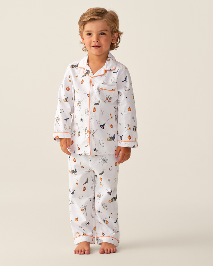 A young child with light brown hair stands barefoot, wearing the Kid’s Twill Pajama Set in Trick or Treat. The white pajamas feature whimsical animal and plant patterns, accented with orange trim along the collar, cuffs, and pocket. The background is plain and light.