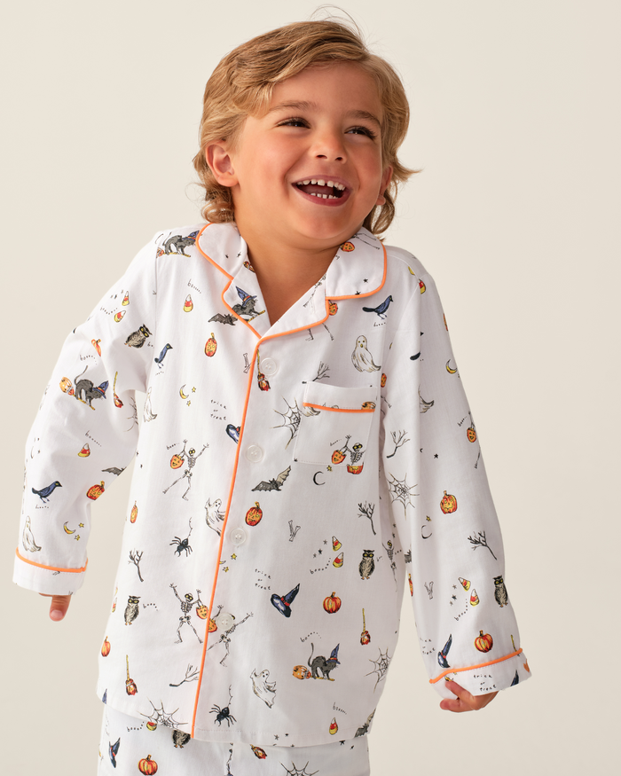 A smiling child with light brown hair wears the Kid’s Twill Pajama Set in Trick or Treat, featuring white pajamas decorated with Halloween-themed illustrations such as pumpkins, ghosts, cats, and bats, set against a plain light background.