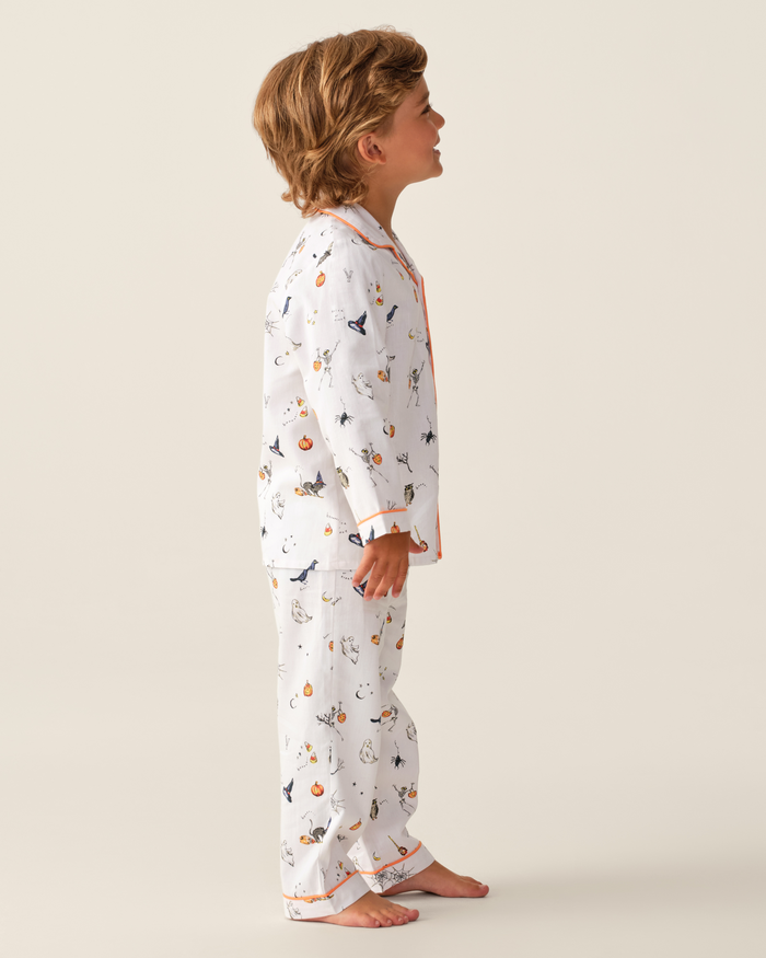 A young child stands barefoot in profile, wearing the Kid’s Twill Pajama Set in Trick or Treat, featuring playful animal and pumpkin prints with vibrant orange trim accents. The backdrop is plain and light-colored.