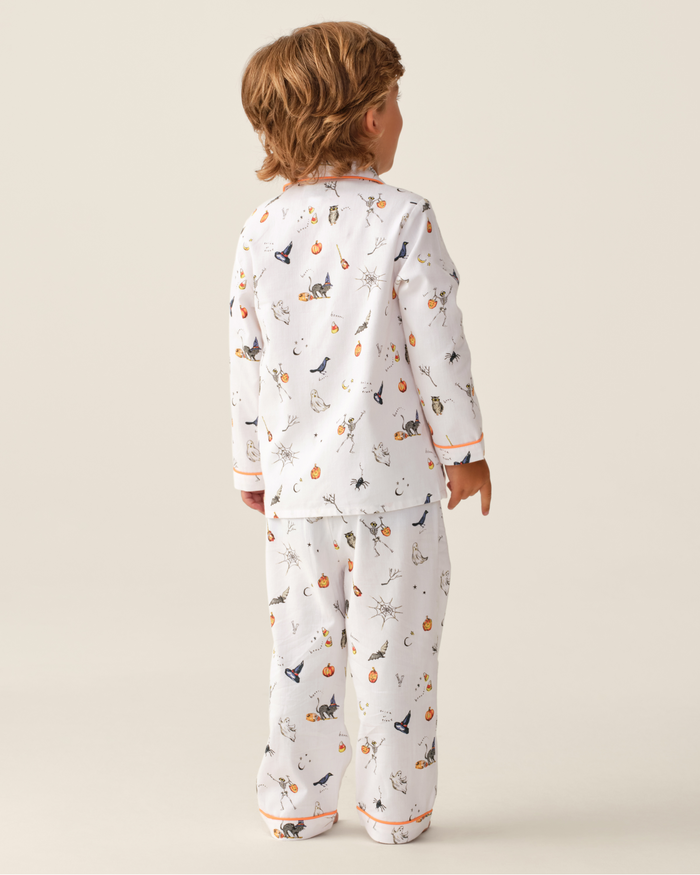 A young child stands facing away, wearing the Kid’s Twill Pajama Set in Trick or Treat—white pajamas decorated with festive Halloween prints like pumpkins, cats, and bats. The background is plain and light-colored.