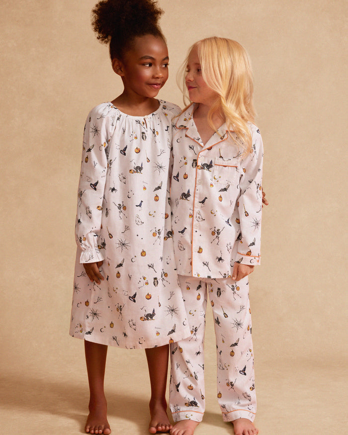 Petite Plumes Kids Twill Pajama Set in Trick or Treat features whimsical white pajamas adorned with Halloween-themed pumpkins, spiders, moons, and playful animals. This luxury sleepwear includes a button-up top with orange trim and matching pants in a festive print.
