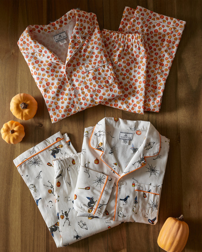Two sets of Petite Plume Kids Twill Pajama Sets rest on a wooden surface—one with pumpkins, the other featuring Trick or Treat prints with bats and spiders. Surrounded by small pumpkins, this luxury sleepwear adds a festive touch to your bedtime routine.
.