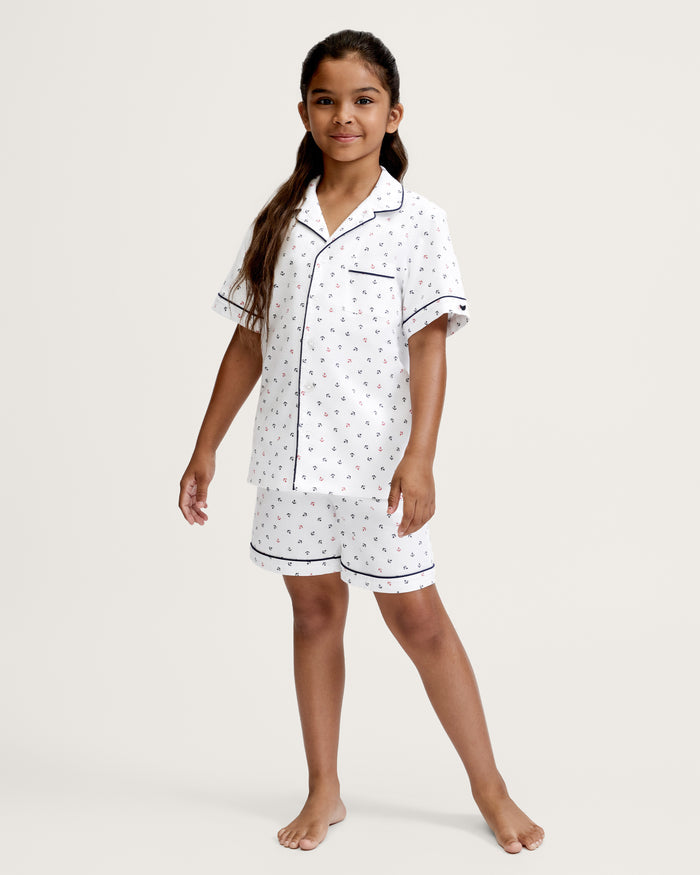 A young girl stands barefoot against a plain light background, wearing the Kid’s Twill Pajama Short Set in Anchors Away White, which features a collared, short-sleeved top with a front pocket and small colorful anchor patterns.