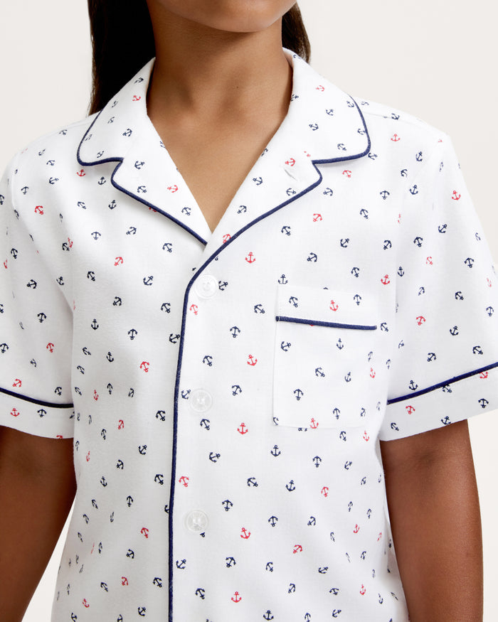 A child is wearing the Kid’s Twill Pajama Short Set in Anchors Away White, featuring a white pajama top with small navy and red anchor prints, navy piping on the collar, pocket, and sleeves, and white buttons.