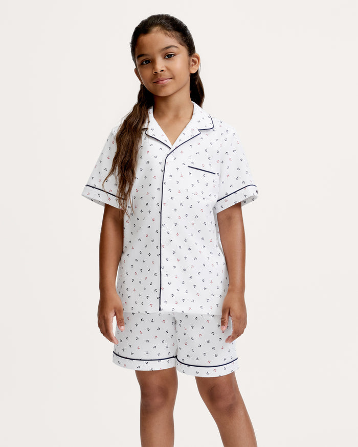 A young girl with long hair wears the Kid’s Twill Pajama Short Set in Anchors Away White, featuring white short-sleeve, button-up pajamas with a small anchor print and navy piping, standing against a plain light background.