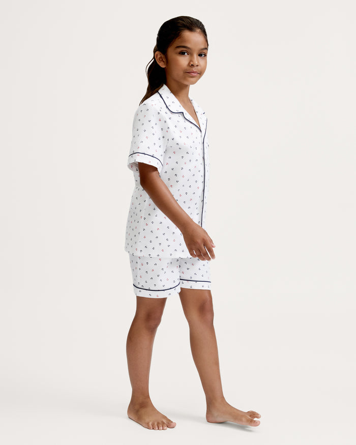 A young girl stands barefoot, wearing the Kid’s Twill Pajama Short Set in Anchors Away White, which features short sleeves, a button-up front, a subtle pattern of small anchors, and navy piping. She faces slightly to the side against a plain white background.