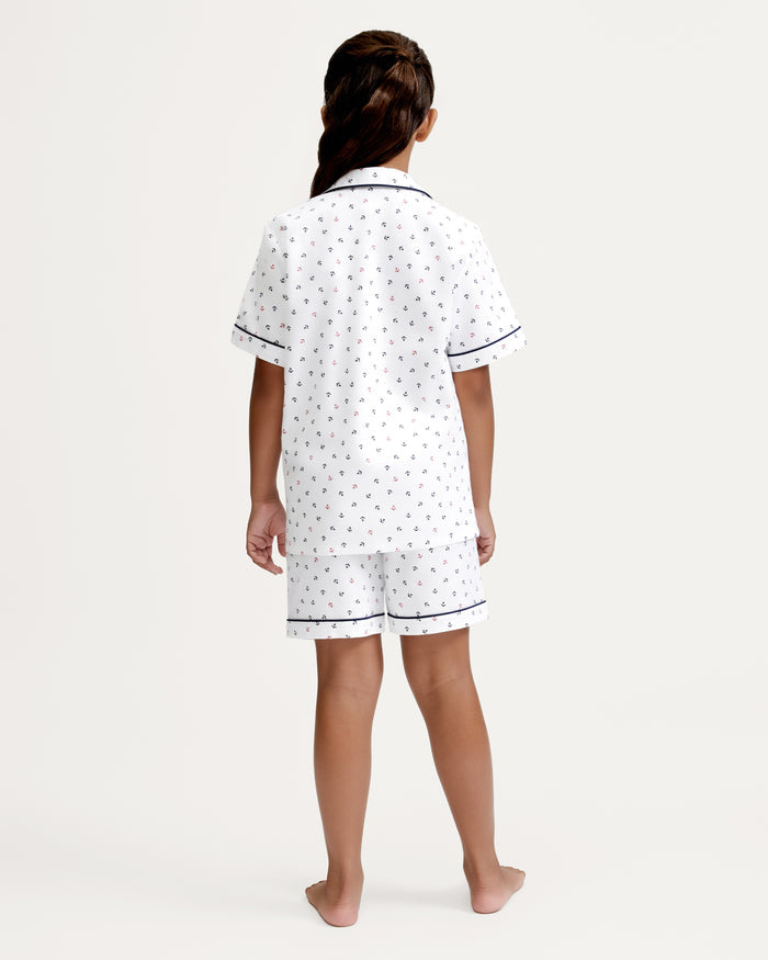 A child with long brown hair stands barefoot facing away, wearing the Kid’s Twill Pajama Short Set in Anchors Away White, featuring white short-sleeved pajamas with a small print and navy trim, against a plain white background.
