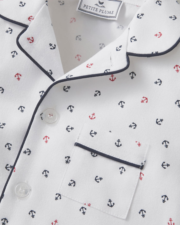 Close-up of the Kid’s Twill Pajama Short Set in Anchors Away White, showing white pajamas with a navy piped collar and pocket, decorated with a pattern of small red and navy anchors. A Petite Plume label is visible inside the collar.