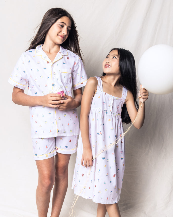 Two girls in the Kids Twill Pajama Short Set in Birthday Wishes by Petite Plume smile at each other. One holds a cupcake, the other a white balloon, against a light background, capturing the dreamy essence of playful celebrations with luxurious sleepwear and pastel fantasy prints.