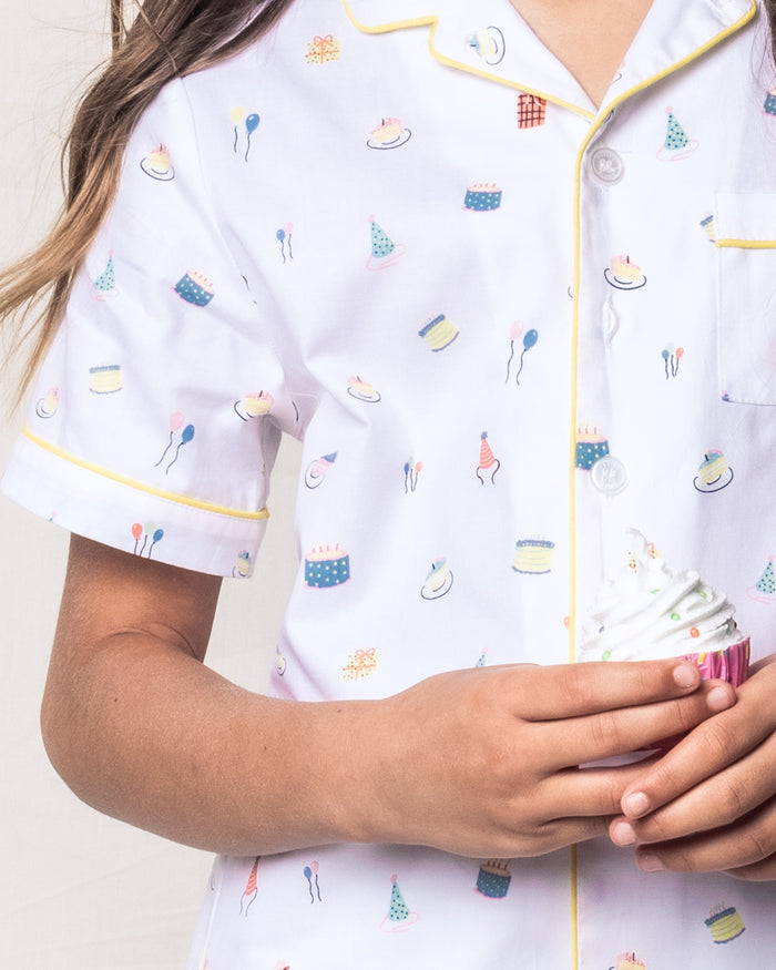 A child in Petite Plumes Kids Twill Pajama Short Set in Birthday Wishes, featuring a whimsical cake and balloon print with yellow piping and buttons, holds a cupcake with white frosting while brown hair peeks through.