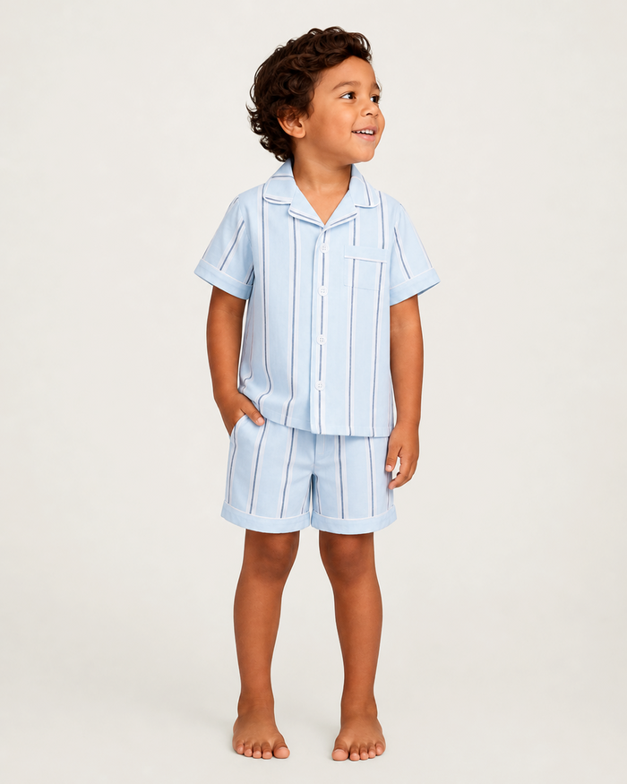 A young child stands barefoot, smiling, with one hand in their pocket, wearing the Kid’s Twill Pajama Short Set in Coastal Stripe—a light blue matching pajama set with short sleeves and shorts featuring white and navy vertical stripes.