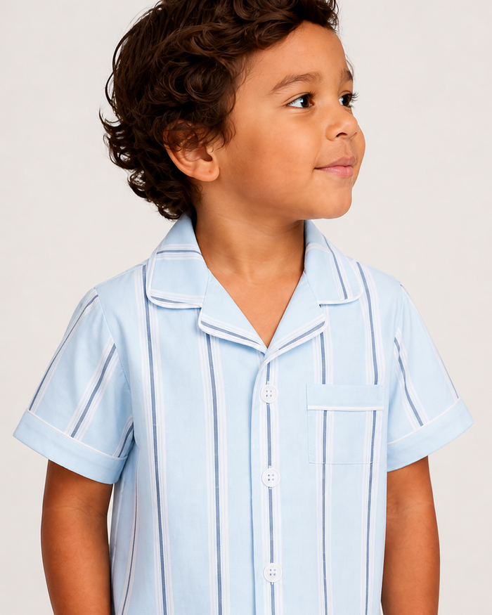 A young child with curly brown hair wears the Kid’s Twill Pajama Short Set in Coastal Stripe—a light blue, short-sleeve button-up featuring white and blue vertical stripes—and smiles slightly while looking to the side against a plain background.