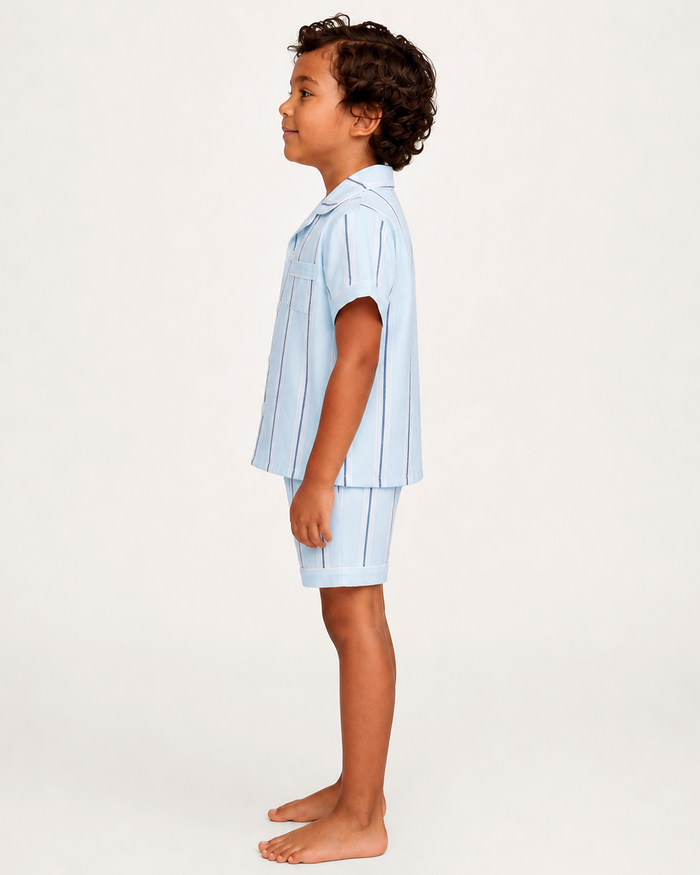 A young child with curly hair stands in profile, barefoot, wearing the Kid’s Twill Pajama Short Set in Coastal Stripe—a light blue, short-sleeve set with vertical stripes. The background is plain and light-colored.