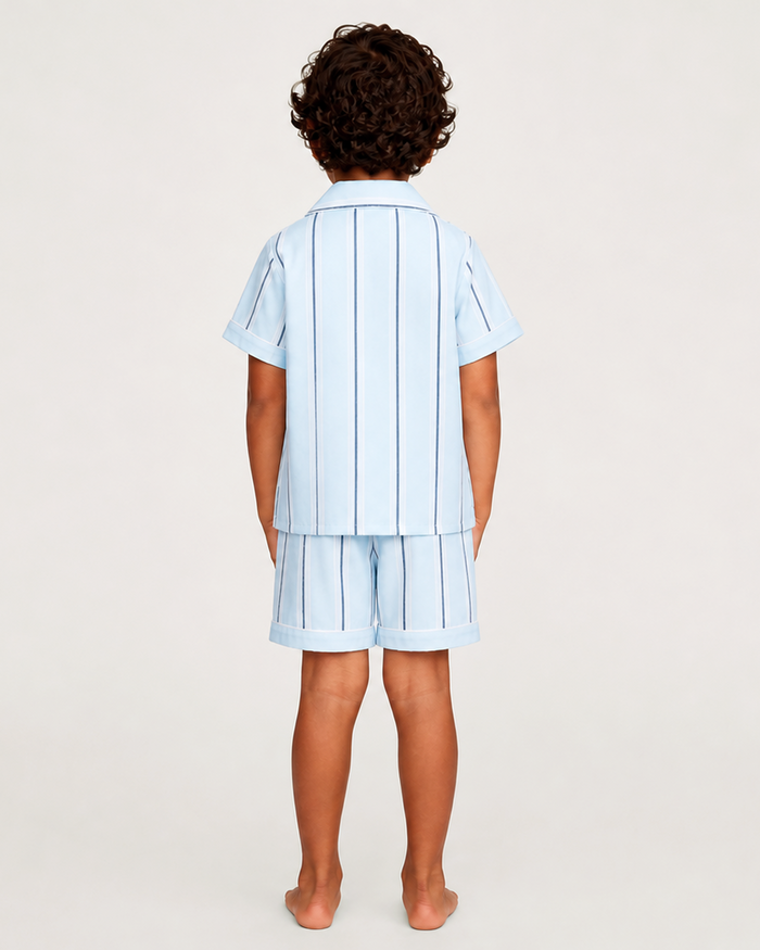 A child with curly hair stands barefoot, facing away, wearing the Kid’s Twill Pajama Short Set in Coastal Stripe—a light blue short-sleeve pajama top with dark vertical stripes and matching shorts—against a plain background.