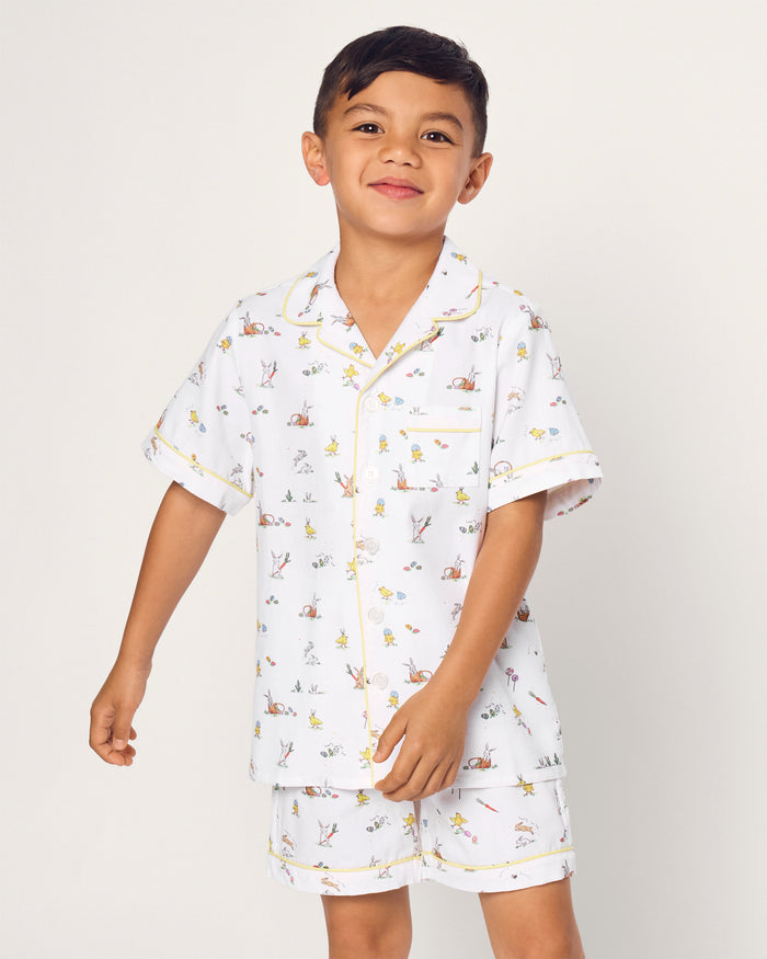Two smiling children in luxurious Petite Plume sleepwear sit on the floor. They sport matching Kids Twill Short Sets in an Easter Frolic print. One child cuddles a stuffed bunny, embodying comfort and joy against a plain backdrop.