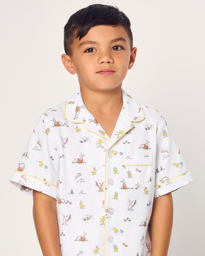 Two smiling children in luxurious Petite Plume sleepwear sit on the floor. They sport matching Kids Twill Short Sets in an Easter Frolic print. One child cuddles a stuffed bunny, embodying comfort and joy against a plain backdrop.