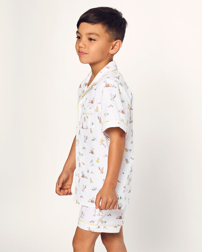 Two smiling children in luxurious Petite Plume sleepwear sit on the floor. They sport matching Kids Twill Short Sets in an Easter Frolic print. One child cuddles a stuffed bunny, embodying comfort and joy against a plain backdrop.