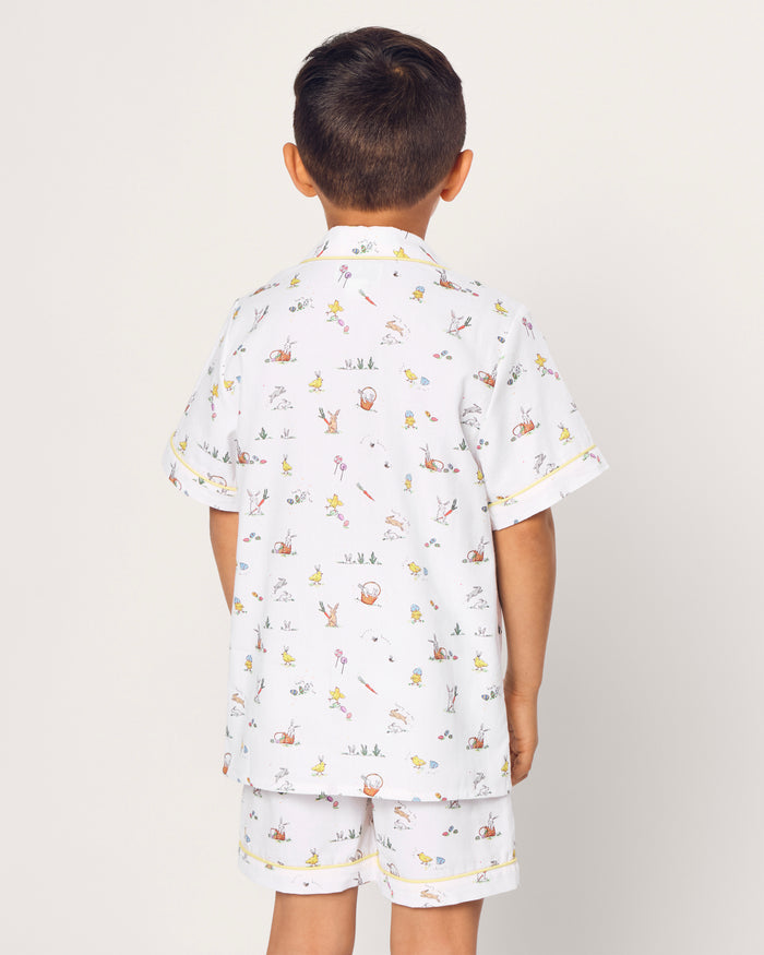 Two smiling children in luxurious Petite Plume sleepwear sit on the floor. They sport matching Kids Twill Short Sets in an Easter Frolic print. One child cuddles a stuffed bunny, embodying comfort and joy against a plain backdrop.