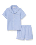 A young boy smiles in Petite Plumes Kids Twill Pajama Short Set, featuring short sleeves and a collar. Made from lightweight cotton, the French Blue Seersucker set includes a chest pocket and matching shorts. He stands against a plain light beige background.