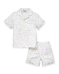 A close-up of a child in the Kid's Twill Pajama Short Set in Good Luck Charms, featuring white fabric with light green trim and pastel rainbows, horseshoes, four-leaf clovers, and stars. Only the upper torso is shown.
