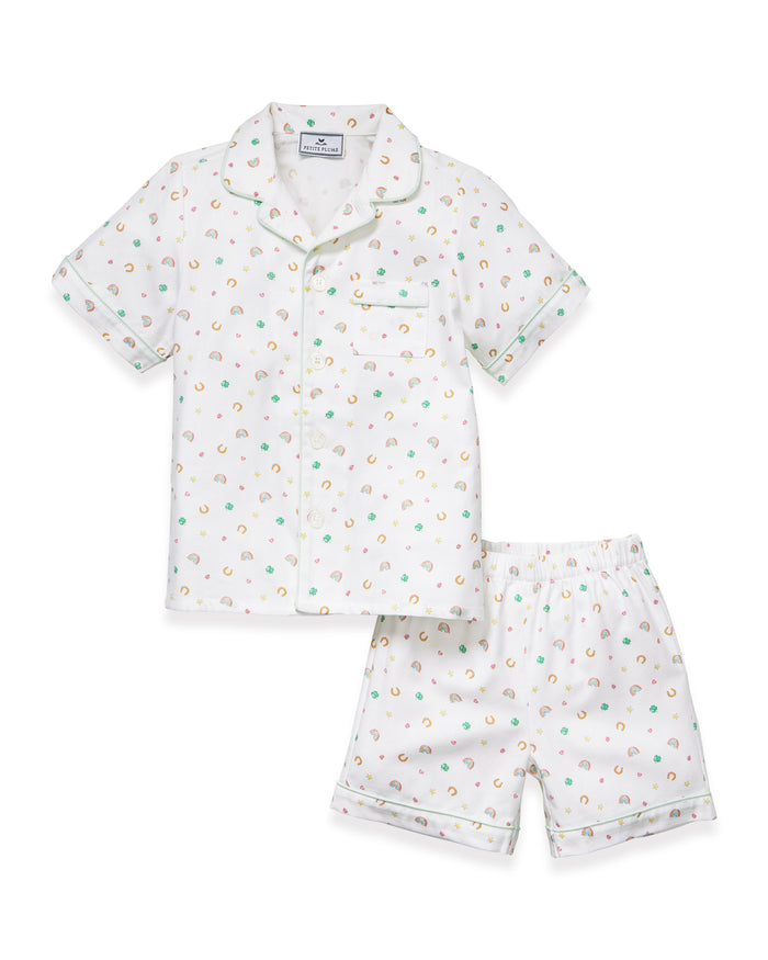 A close-up of a child in the Kid's Twill Pajama Short Set in Good Luck Charms, featuring white fabric with light green trim and pastel rainbows, horseshoes, four-leaf clovers, and stars. Only the upper torso is shown.