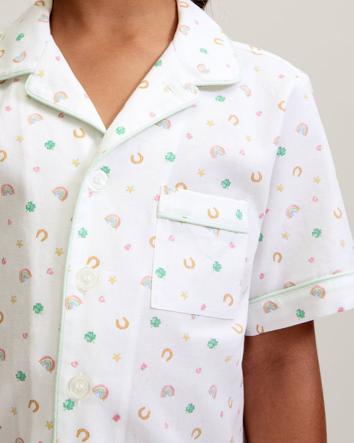 A close-up of a child in the Kid's Twill Pajama Short Set in Good Luck Charms, featuring white fabric with light green trim and pastel rainbows, horseshoes, four-leaf clovers, and stars. Only the upper torso is shown.