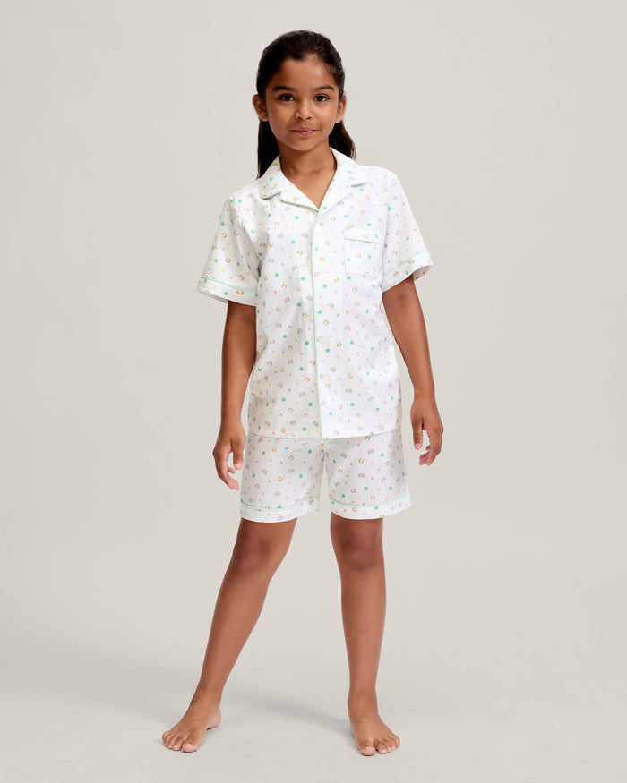 A close-up of a child in the Kid's Twill Pajama Short Set in Good Luck Charms, featuring white fabric with light green trim and pastel rainbows, horseshoes, four-leaf clovers, and stars. Only the upper torso is shown.