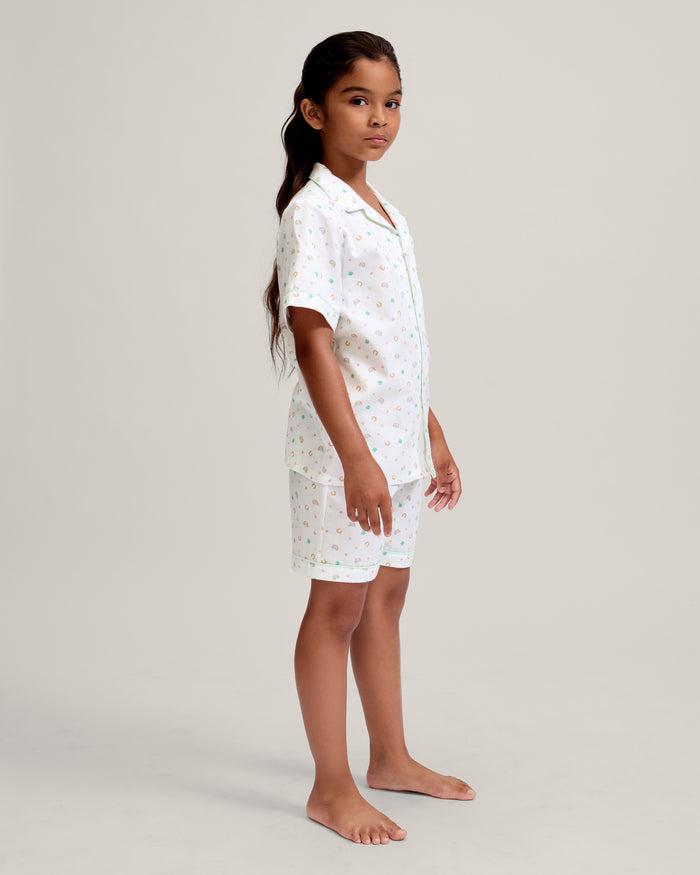 A close-up of a child in the Kid's Twill Pajama Short Set in Good Luck Charms, featuring white fabric with light green trim and pastel rainbows, horseshoes, four-leaf clovers, and stars. Only the upper torso is shown.