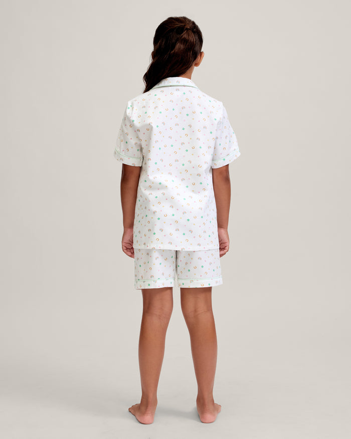 A close-up of a child in the Kid's Twill Pajama Short Set in Good Luck Charms, featuring white fabric with light green trim and pastel rainbows, horseshoes, four-leaf clovers, and stars. Only the upper torso is shown.