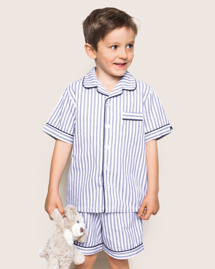 A young boy beams in his luxurious navy and white Kids Twill Pajama Short Set by Petite Plume, crafted from yarn-dyed cotton. Holding a plush teddy bear, he stands against a simple, light beige background.