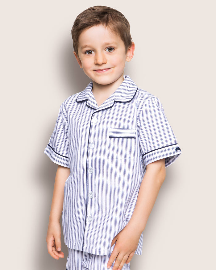 A young boy smiles in Petite Plumes Kids Twill Pajama Short Set in Navy French Ticking. He stands against a plain, light backdrop with short dark hair, hands by his sides, exuding luxurious sleepwear comfort.