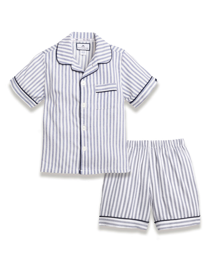 The Kids Twill Pajama Short Set in Navy French Ticking by Petite Plume features a short-sleeve button-up shirt and shorts, crafted from yarn-dyed cotton with vertical blue and white stripes. It includes contrasting piping, a notched collar, a chest pocket, and elastic waistband for ultimate comfort.