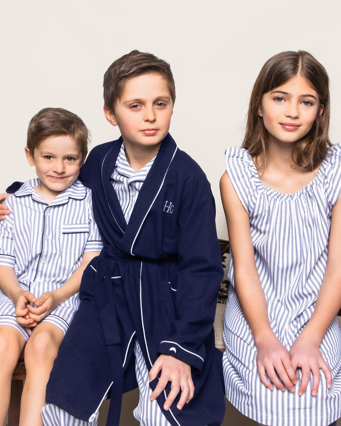 Three children sit together in matching Petite Plume sleepwear, crafted from luxurious yarn-dyed cotton. Two boys on the left wear pajamas and a robe, while a girl on the right wears a striped nightgown. They smile warmly at the camera against a plain backdrop.