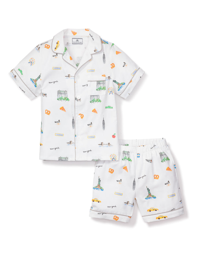 Petite Plumes Kids Twill Pajama Short Set, New York! New York!, offers luxury cotton sleepwear with a short-sleeved button-up shirt and shorts. It features illustrations of NYC landmarks and motifs such as the Statue of Liberty, Empire State Building, hot dogs, and taxis.