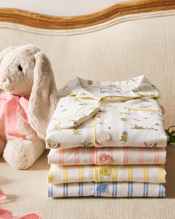 A plush white bunny with a pink ribbon sits next to a neatly folded Petite Plume Kids Twill Pajama Short Set in Periwinkle and White Stripe on a beige upholstered couch.