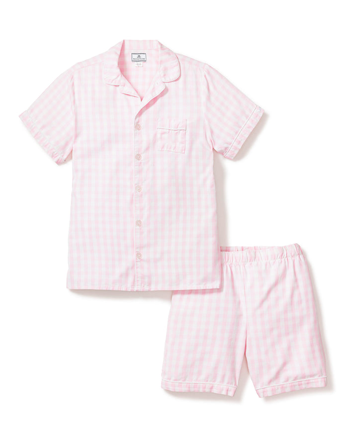 Three folded Petite Plume Kids Twill Pajama Short Sets in pink gingham rest on a textured surface, featuring charming designs of pink hearts, floral toile patterns, and pink stripes. Crafted from yarn-dyed cotton, they exude quality. Elegantly surrounded by white flowers for enhanced presentation.
