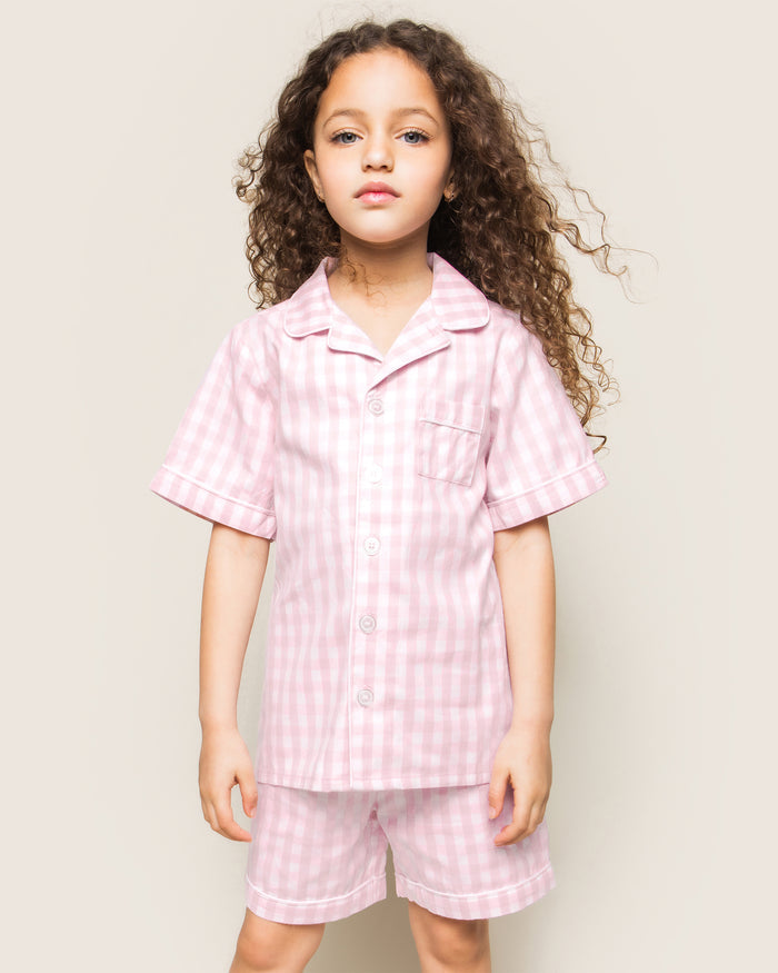 Three folded Petite Plume Kids Twill Pajama Short Sets in pink gingham rest on a textured surface, featuring charming designs of pink hearts, floral toile patterns, and pink stripes. Crafted from yarn-dyed cotton, they exude quality. Elegantly surrounded by white flowers for enhanced presentation.
