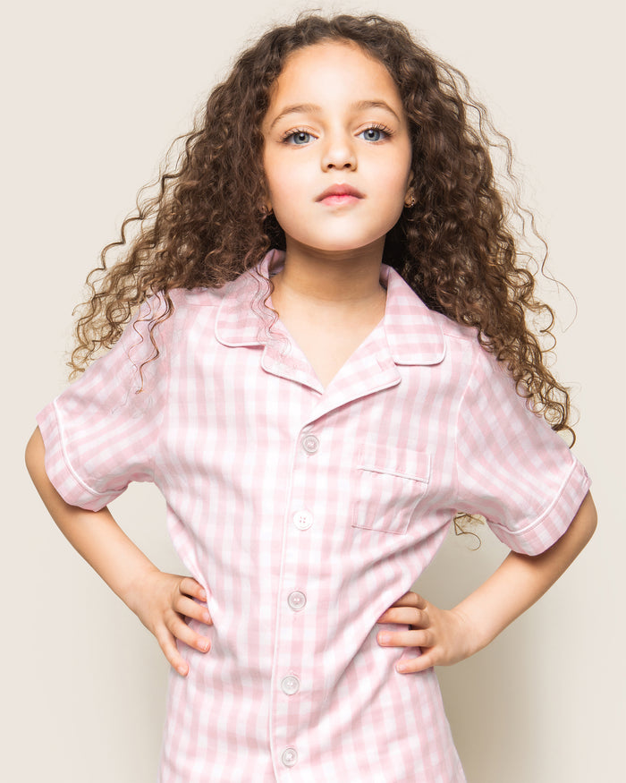 Three folded Petite Plume Kids Twill Pajama Short Sets in pink gingham rest on a textured surface, featuring charming designs of pink hearts, floral toile patterns, and pink stripes. Crafted from yarn-dyed cotton, they exude quality. Elegantly surrounded by white flowers for enhanced presentation.
