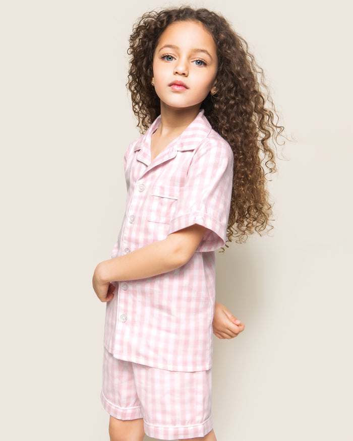 Three folded Petite Plume Kids Twill Pajama Short Sets in pink gingham rest on a textured surface, featuring charming designs of pink hearts, floral toile patterns, and pink stripes. Crafted from yarn-dyed cotton, they exude quality. Elegantly surrounded by white flowers for enhanced presentation.