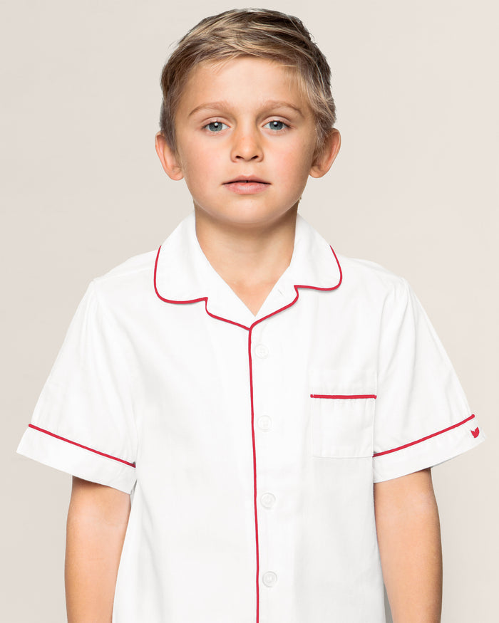 A young boy models Petite Plumes Kids White Twill Short Set with Red Piping. The yarn-dyed cotton set features a button-down shirt with red piping and a pocket on the left side. His short, light-colored hair complements his direct gaze against the plain backdrop.