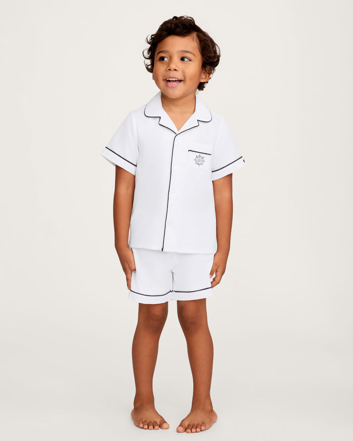 A young child stands barefoot, smiling and looking slightly to the side against a plain background, dressed in the Kid’s Twill Pajama Short Set in White with Yacht Club Embroidery—featuring white short sleeves, black piping, and a small embroidered yacht club design on the pocket.