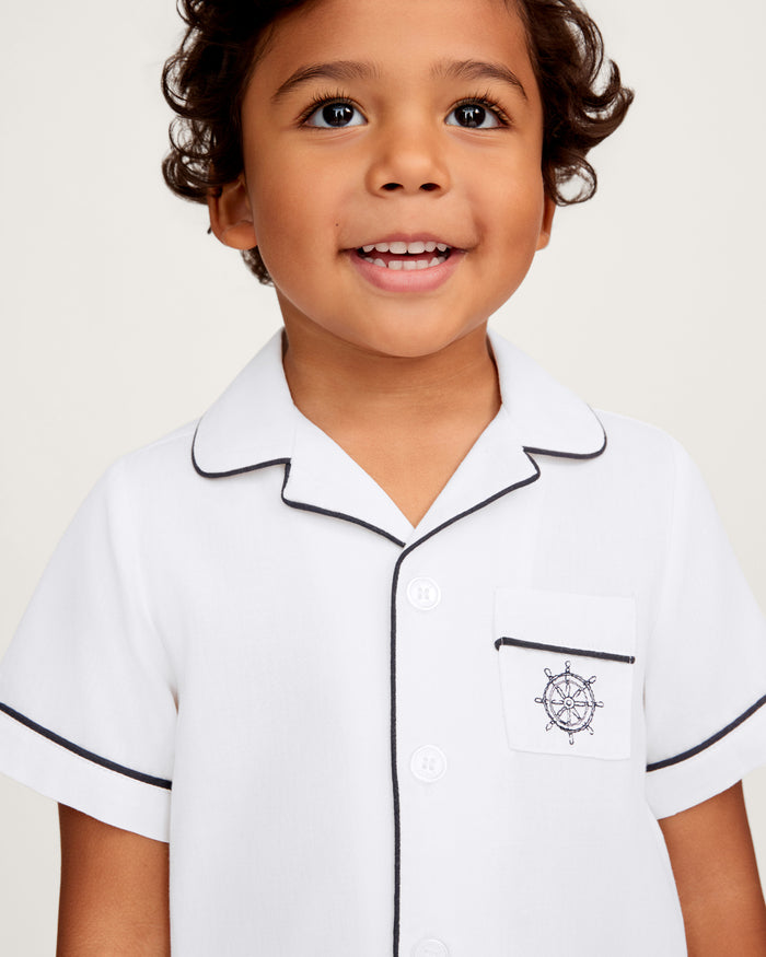 A smiling young child with curly hair wears the Kid’s Twill Pajama Short Set in White, featuring yacht club embroidery and dark piping, standing against a plain background.