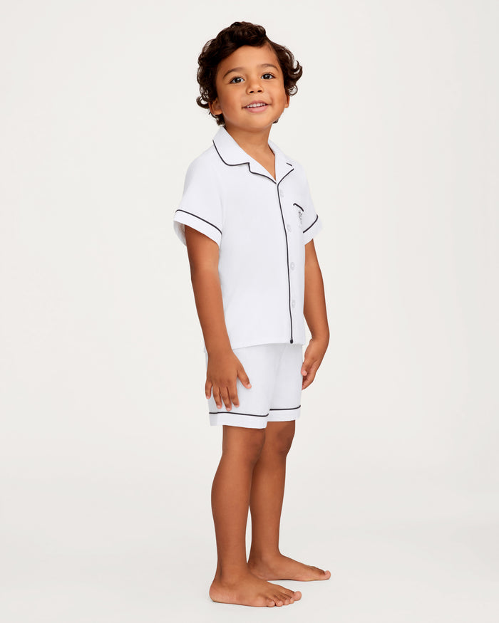A young child with curly hair stands barefoot, smiling while wearing the Kid’s Twill Pajama Short Set in White with Yacht Club Embroidery. The short-sleeve pajama set features black piping, a button-up shirt, and matching shorts. The background is plain and light-colored.